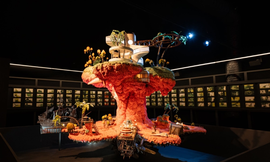 A detailed model from Gorillaz’s House of Kong exhibition, featuring a futuristic white building perched on a vibrant red rock formation, surrounded by miniature palm trees, winding pathways, and set pieces, displayed under dramatic lighting in a dark gallery space.