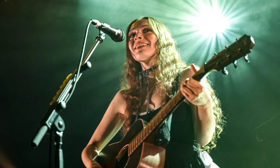 Holly Humberstone performing live on stage, smiling while playing an acoustic guitar under bright stage lights.