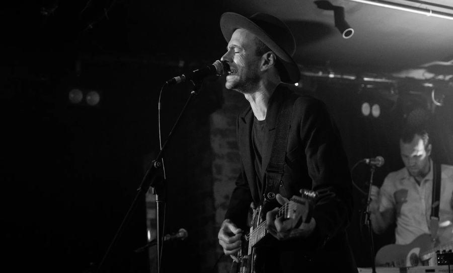 Finn Andrews of The Veils performing live on stage in a dark venue, wearing a hat and playing guitar while singing into a microphone.