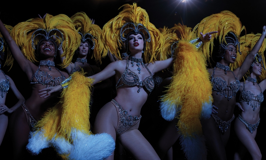 Taylor Swift performs in glittering showgirl costume with feathered headdress, surrounded by dancers in matching outfits for The Life of a Showgirl.