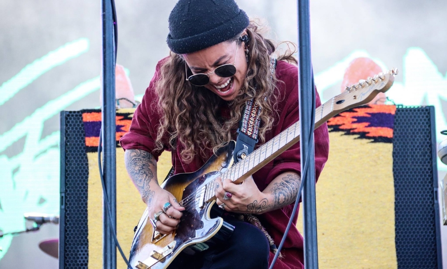 Tash Sultana performs live on stage, wearing sunglasses and a beanie while playing an electric guitar with intensity.