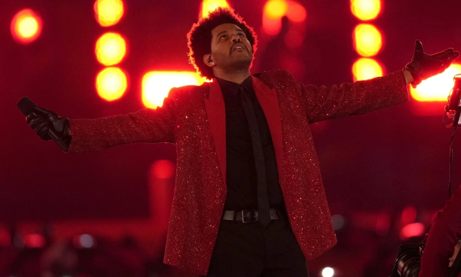 The Weeknd performs in a glittering red jacket during a live show, holding a microphone with arms outstretched against a backdrop of red lights.