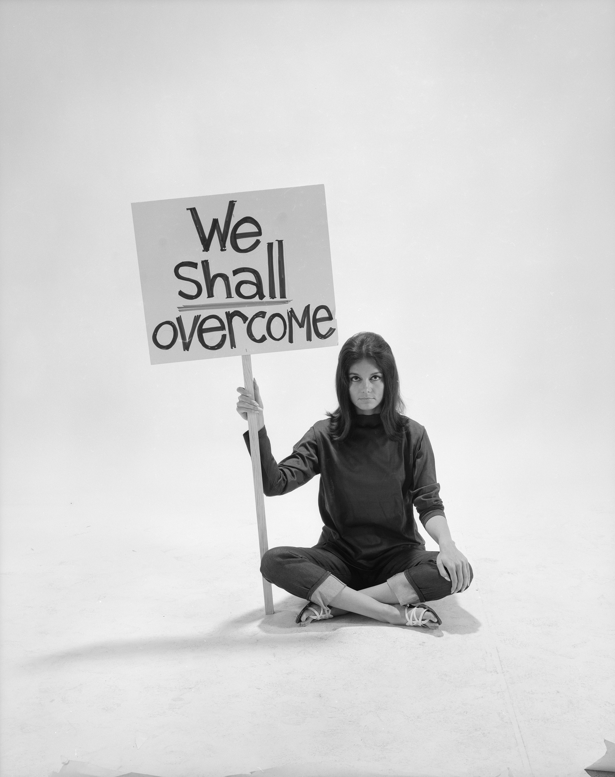 Writer Gloria Steinem sitting cross-legged on floor w. sign "We Shall Overcome" re pop culture